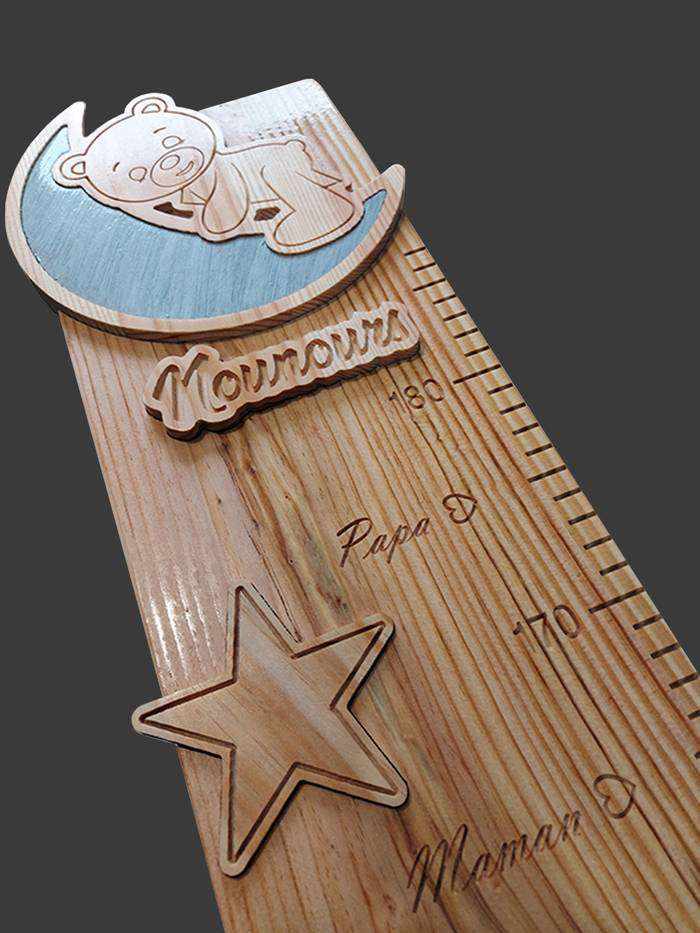 Pour cette toise en bois, le thème est le ciel étoilé. Un ourson sculpté sur son croissant de lune, avec le mot Nounours.