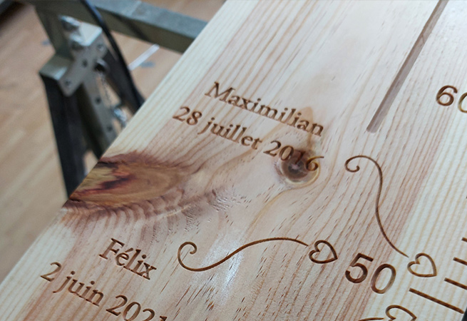 Prénoms et dates de naissance sculptés sur une toise en bois massif, avec une flèche en arabesque et un cœur indiquant la taille à la naissance